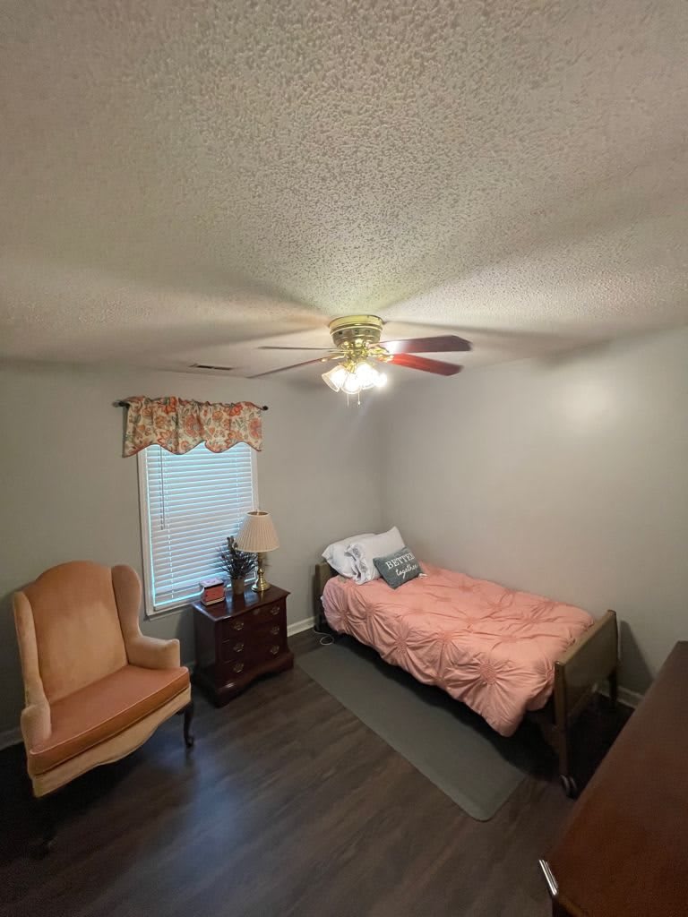 A cozy bedroom with a single bed, armchair, nightstand, and a ceiling fan.