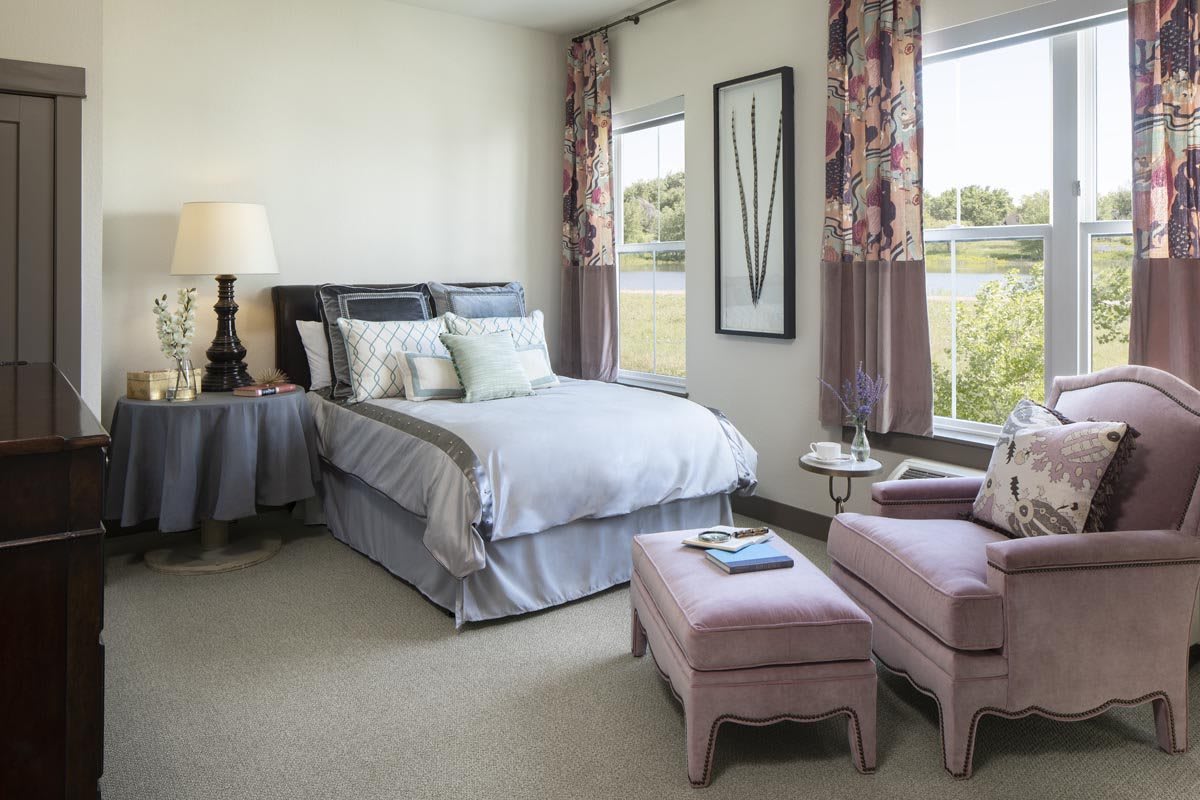 A cozy bedroom featuring a well-made bed, adjacent nightstand with a lamp, and two upholstered armchairs. The room is decorated with floral curtains and art on the wall.