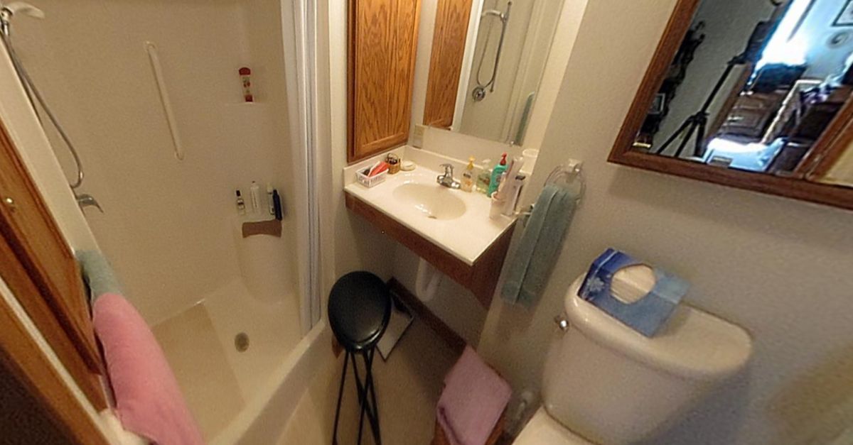 A compact bathroom featuring a sink with toiletries, a mirror, a toilet, a shower area, and a stool.