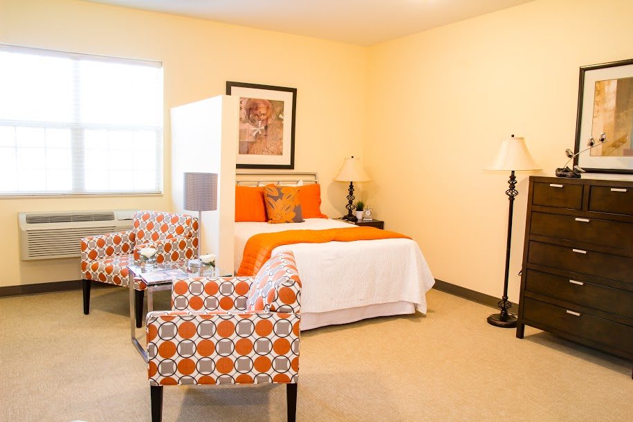 A cozy bedroom with orange accents, featuring a bed, patterned chairs, and stylish decor.