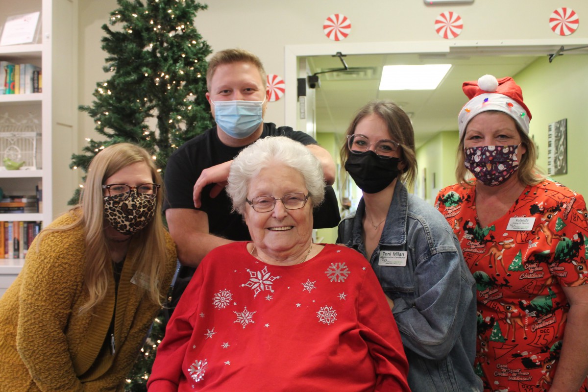 A group of people celebrating Christmas together indoors, with an elderly woman in a red festive sweater at the center.