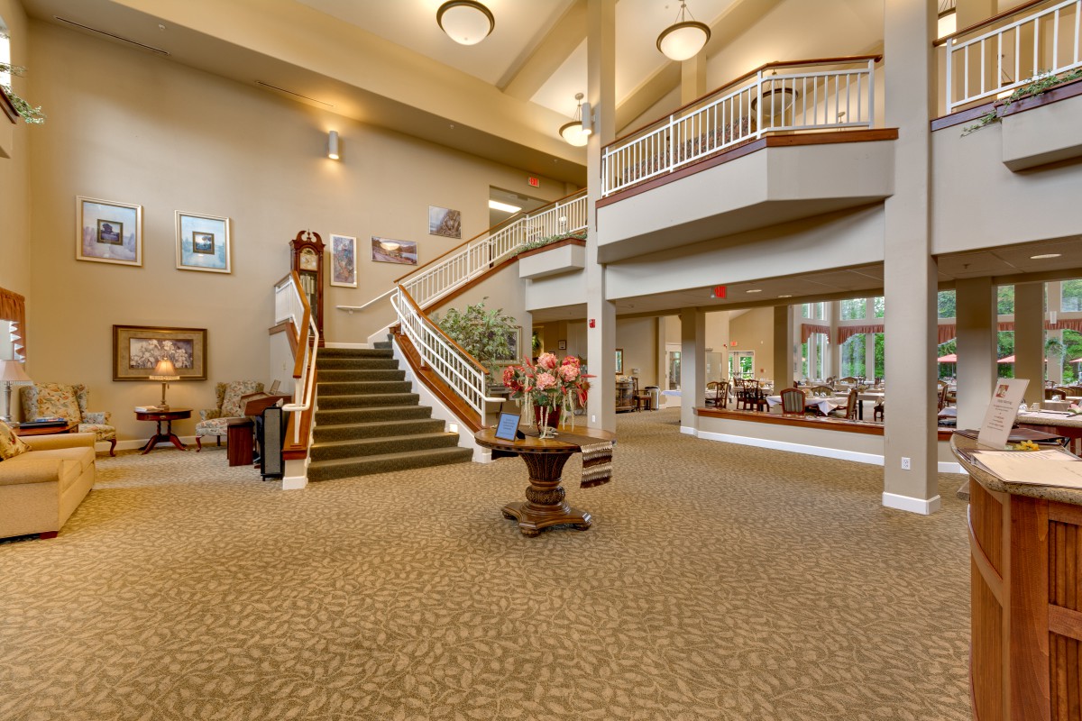 A spacious and well-lit lobby featuring a carpeted floor, central table with flowers, and a grand staircase.