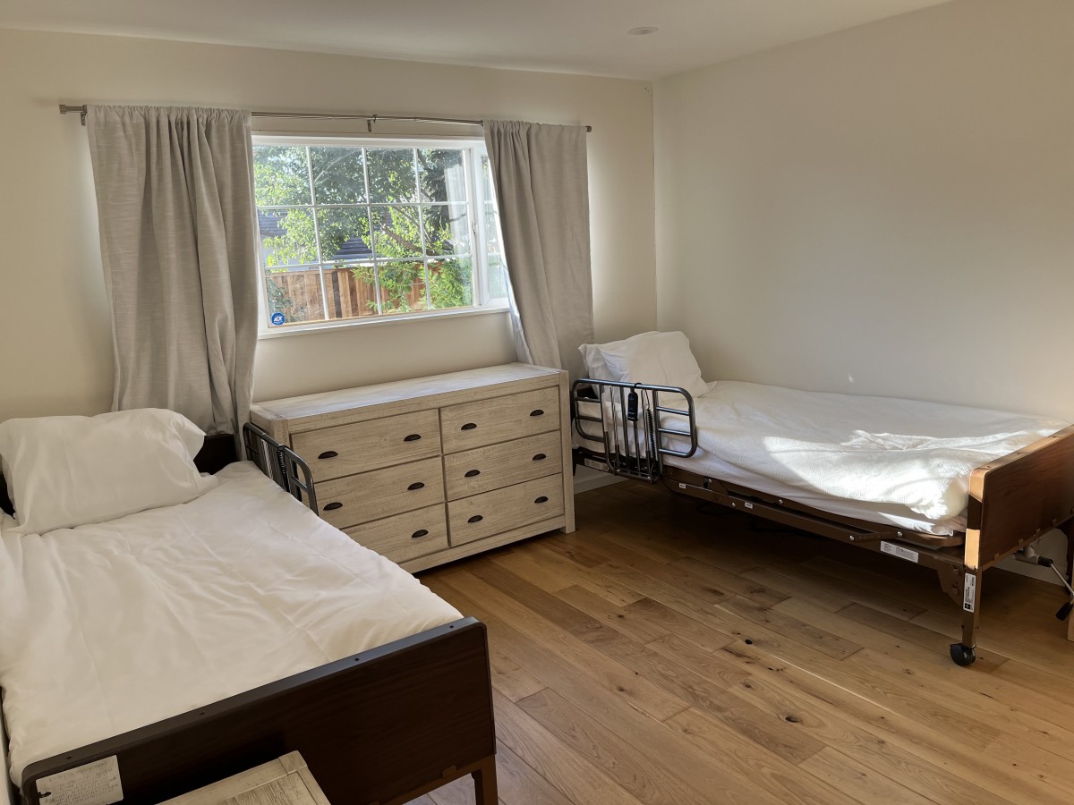 A bright bedroom with two beds, a dresser, and natural light streaming through the window.