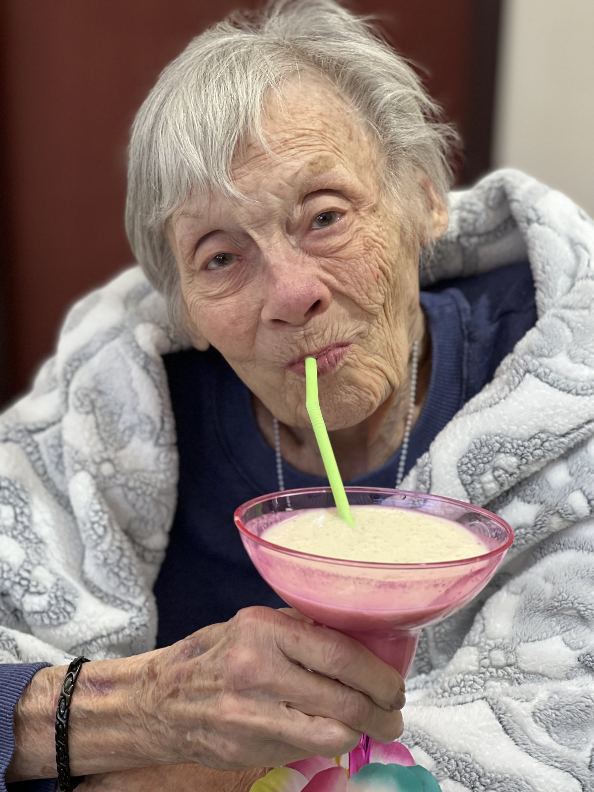 An elderly woman enjoying a milkshake while wrapped in a cozy blanket.
