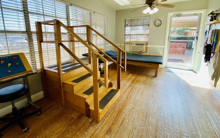 A brightly lit rehabilitation room features a set of wooden stairs and an exercise table.