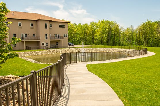 A scenic view of a house by a pond with a pathway and fence, surrounded by a lush lawn and trees on a sunny day.