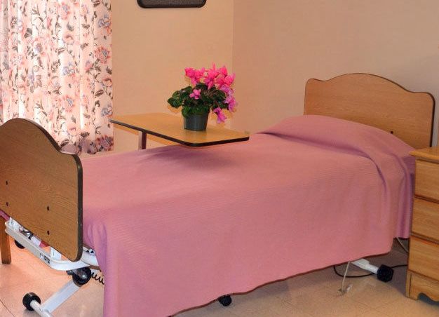 A hospital room with a bed covered in pink bedding, featuring a side table with a flower vase and floral curtains.