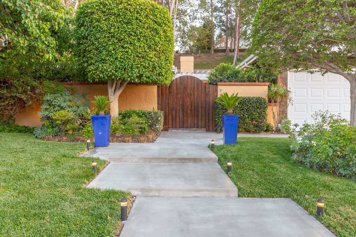 A neatly landscaped garden with a concrete pathway leading to a wooden gate, flanked by tall blue planters and lush greenery.