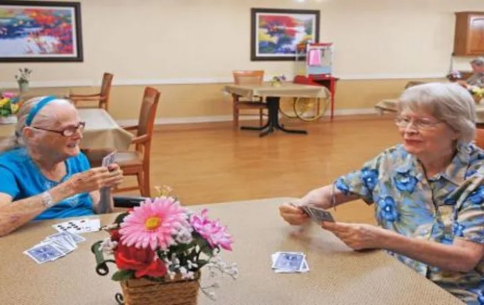 Two elderly individuals playing cards at a table in a communal setting.