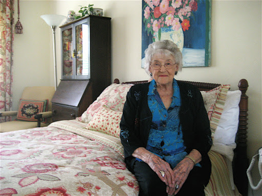 An elderly woman seated in her cozy bedroom, surrounded by warm decor.