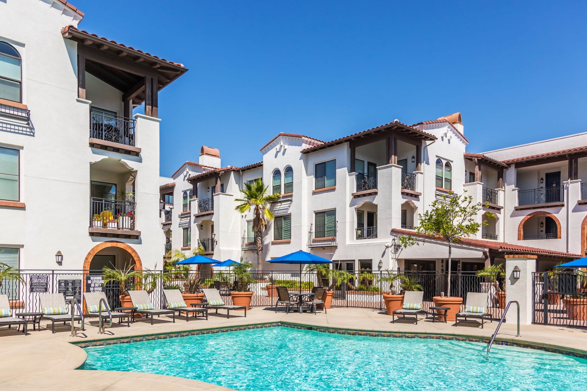 A sunny day at a residential apartment complex featuring a swimming pool surrounded by lounge chairs and umbrellas.
