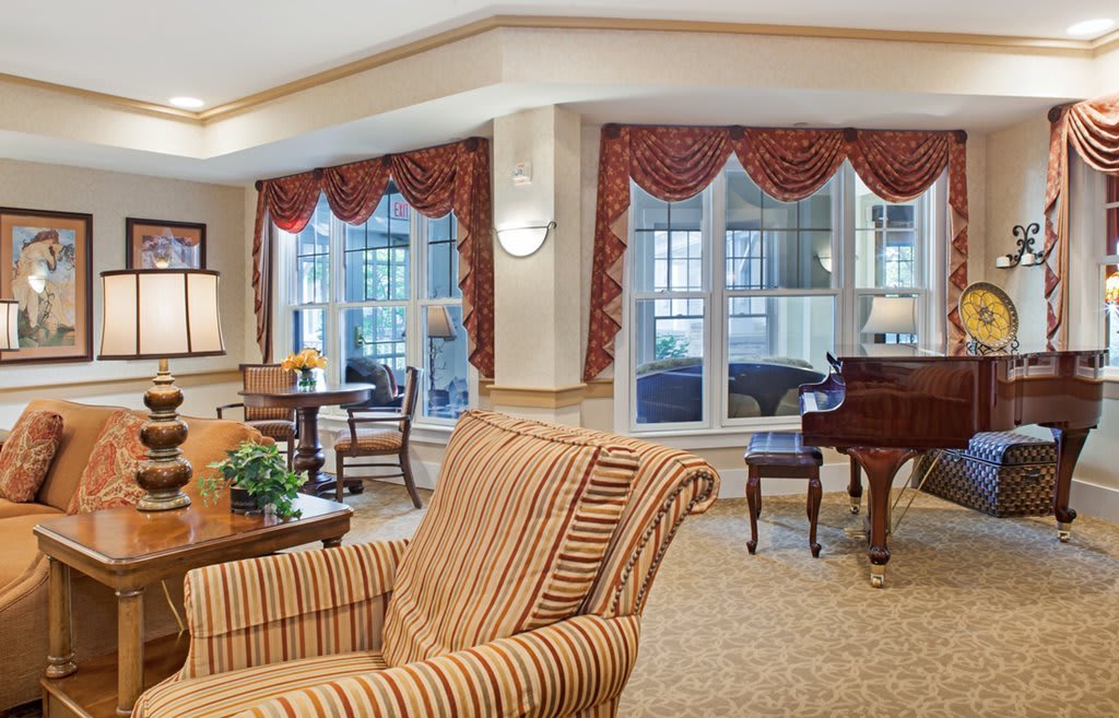 A cozy living room with a piano and comfortable seating, featuring large windows and elegant decor.
