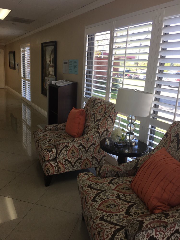 A well-lit lobby area featuring two patterned chairs with orange cushions, a side table with a lamp, and large windows adorned with shutters.