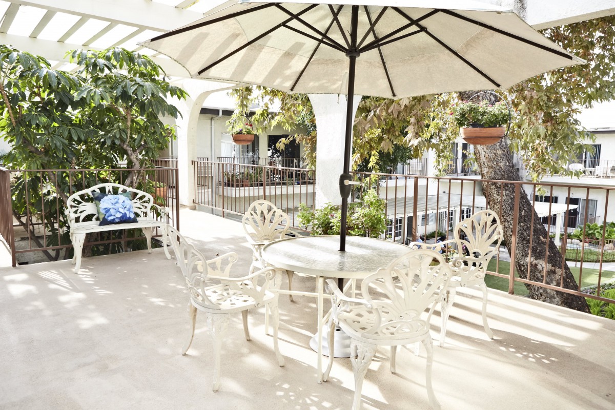 A cozy patio setting with white wrought iron furniture and a large umbrella, surrounded by greenery.