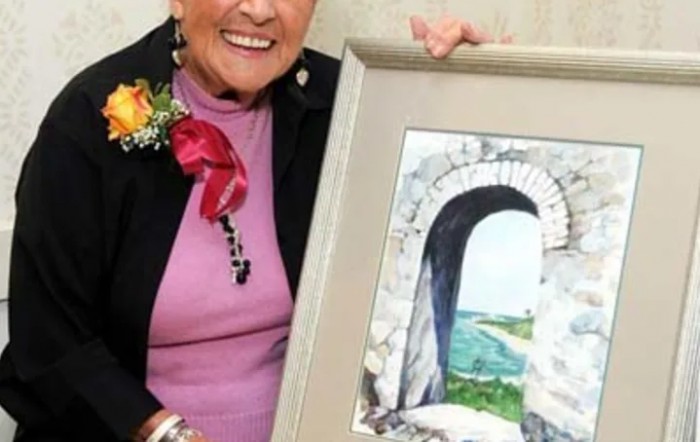 A woman smiles while holding a framed painting of an archway leading to a scenic landscape.