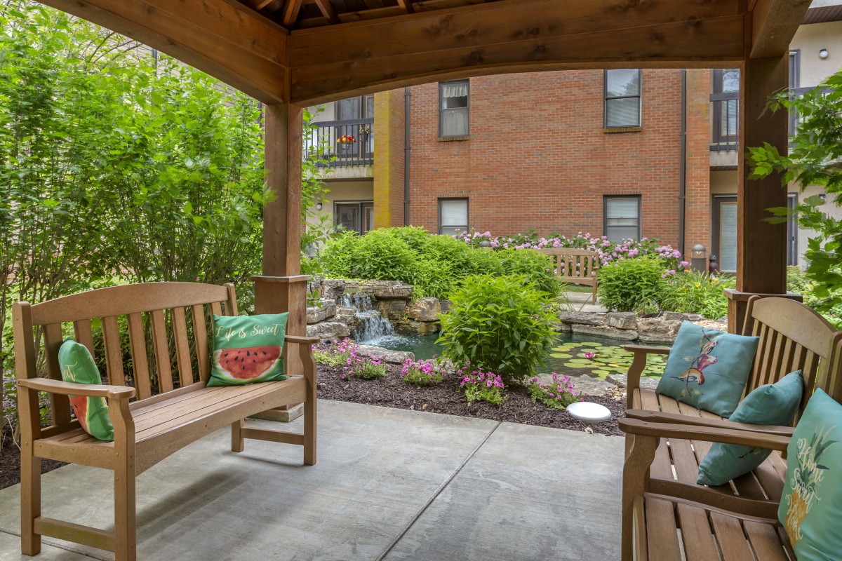 A serene garden view from a wooden gazebo, featuring comfortable benches and vibrant landscaping against the backdrop of a brick building.