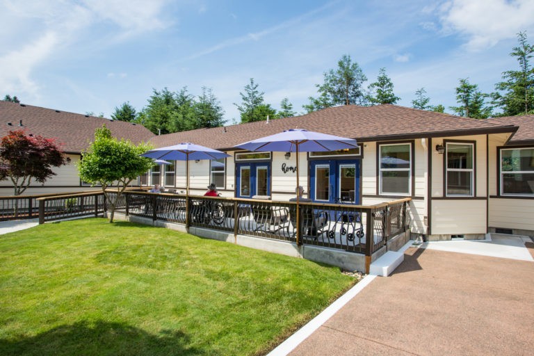 A charming house with a spacious deck, featuring patio furniture and blue umbrellas, set against a well-maintained green lawn.