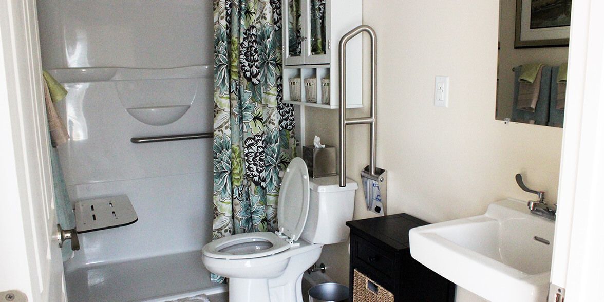 A bathroom with a toilet, shower area with a patterned curtain, sink, and safety grab bar.