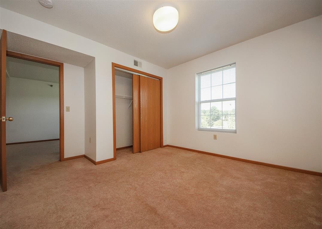 An empty bedroom with a carpeted floor, closet, and a window letting in natural light.