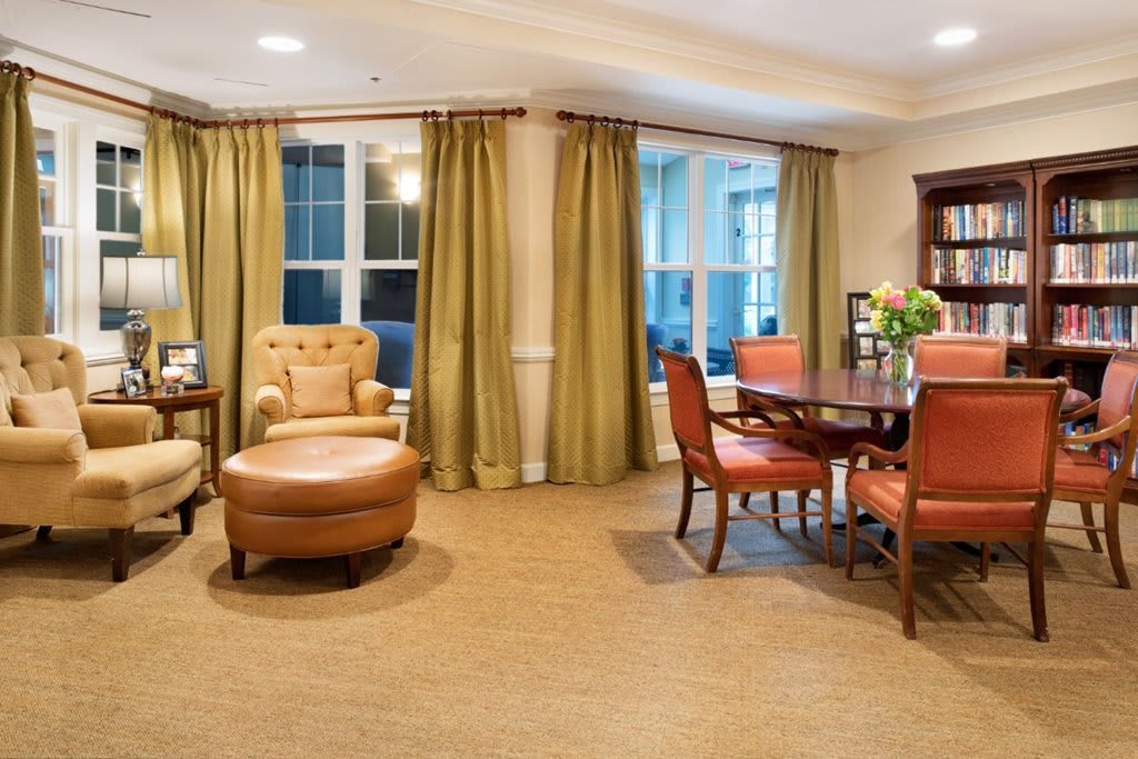 A cozy living and dining area with upholstered chairs, a round dining table, and large windows with green curtains.