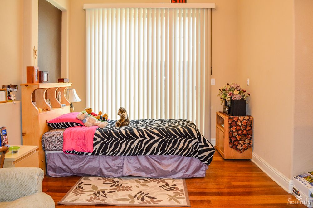 A cozy bedroom with zebra print bedding and a floral rug, featuring wooden floors and vertical blinds.