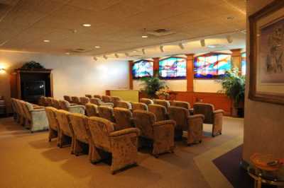 A small auditorium with rows of upholstered chairs facing a wall decorated with stained glass windows and indoor plants.