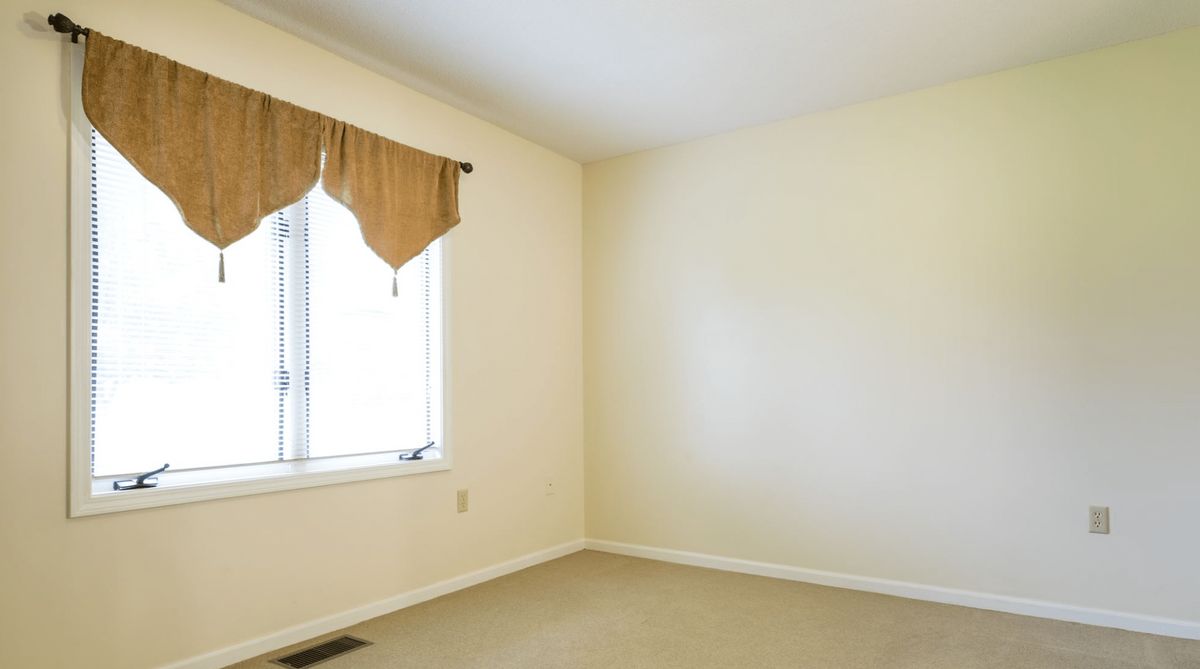 An empty room with cream-colored walls, a window adorned with curtains, and carpeted flooring.