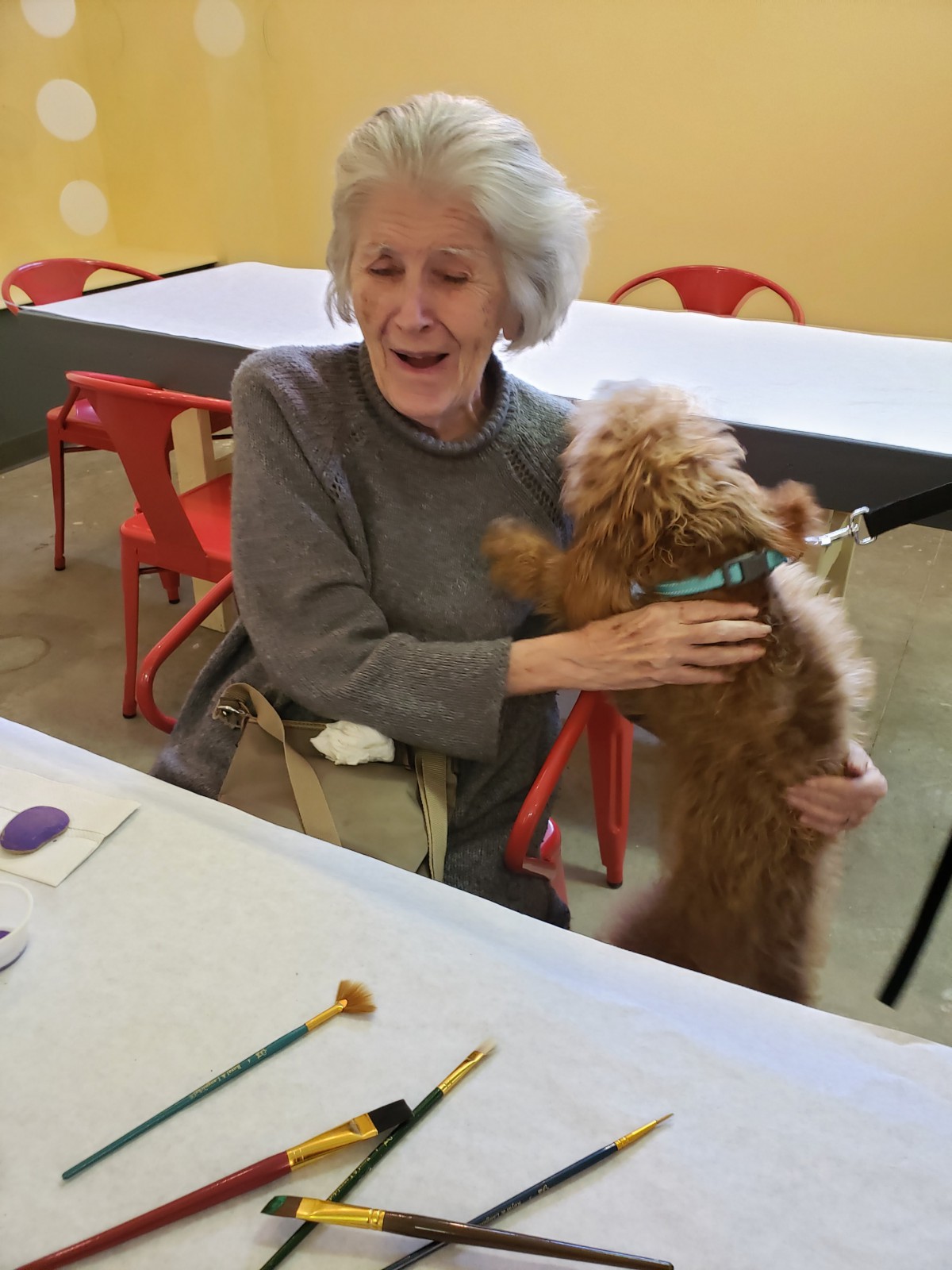 An elderly woman joyfully hugging a dog indoors, with tables and chairs around.