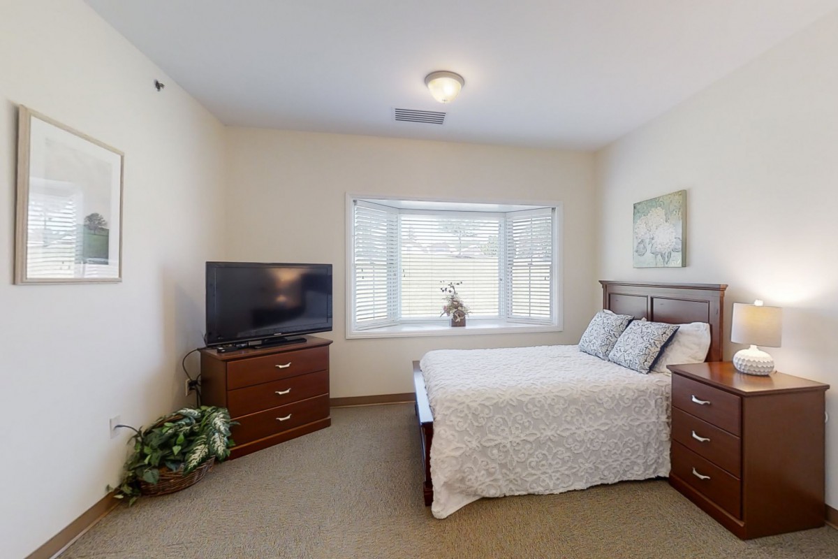 A cozy bedroom with a neatly made bed, a TV on a dresser, and a large window letting in natural light.