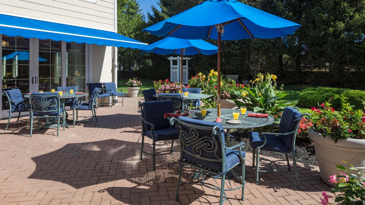A sunny patio with blue umbrellas and outdoor seating, surrounded by a garden.