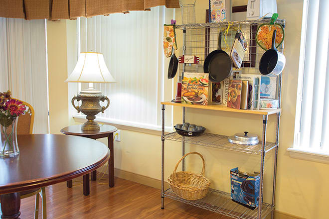 A cozy kitchen corner with a shelf displaying cookbooks and pans, next to a lamp-lit table.