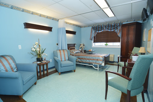 A well-furnished hospital patient room featuring a bed, seating area, and calming blue decor.