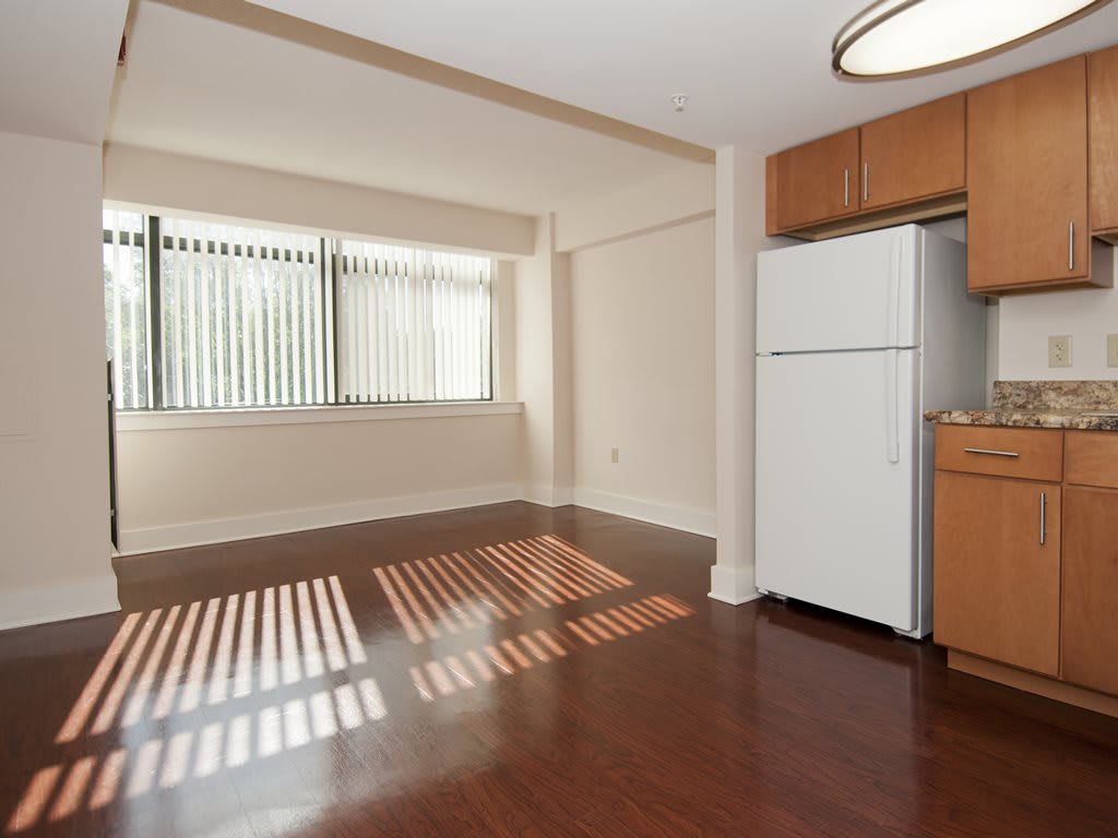 An empty apartment living area with hardwood floors and a kitchen featuring a refrigerator, illuminated by natural light from a large window.