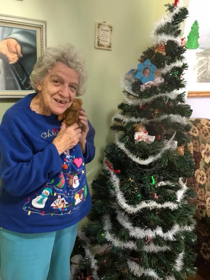 An elderly woman in a festive sweater poses happily beside a decorated Christmas tree.