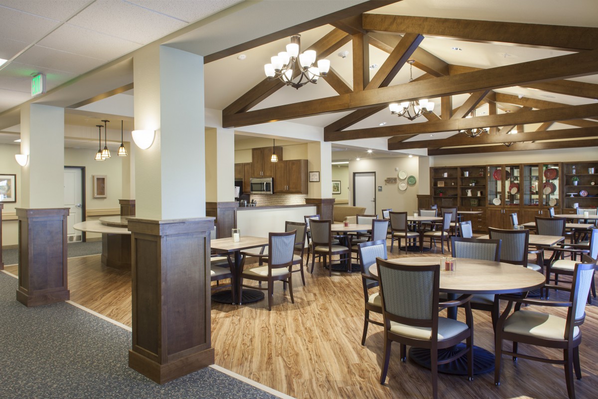A spacious dining area with wooden beams, featuring round tables and chairs, illuminated by elegant chandeliers.