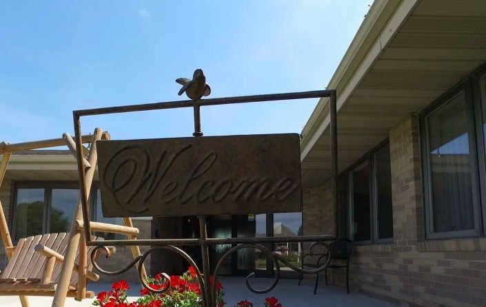 A decorative welcome sign with a bird perched on top, placed near red flowers on a porch.