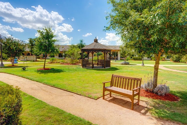 A tranquil park scene featuring a wooden bench along a pathway, with a gazebo and lush greenery.