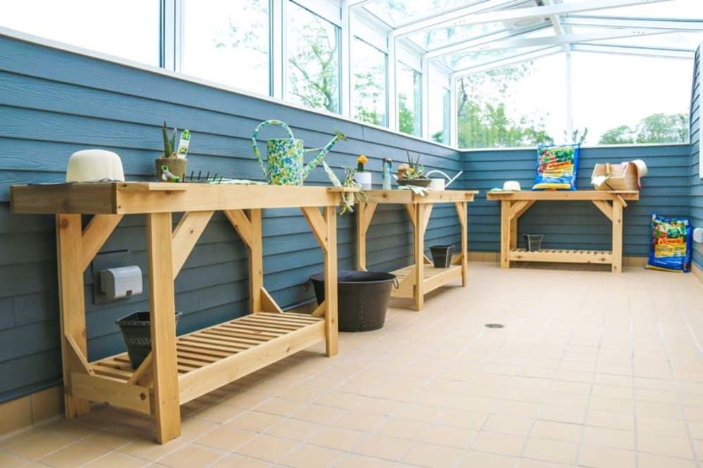 A well-organized greenhouse with wooden potting benches and various gardening tools.