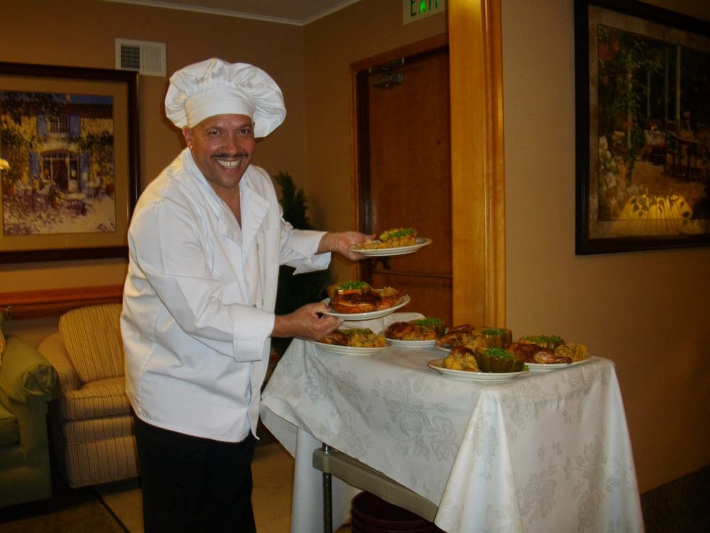 A smiling chef presenting dishes on a table in a kitchen setting.