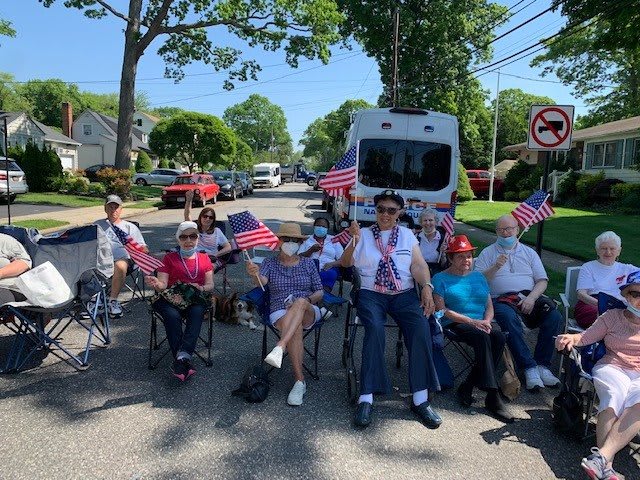 A group of people sitting on chairs along a street, holding American flags during a parade.