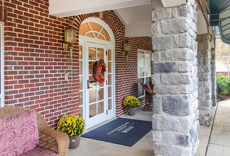 A cozy front porch with a brick wall and glass door adorned with a wreath, featuring potted plants and outdoor seating.