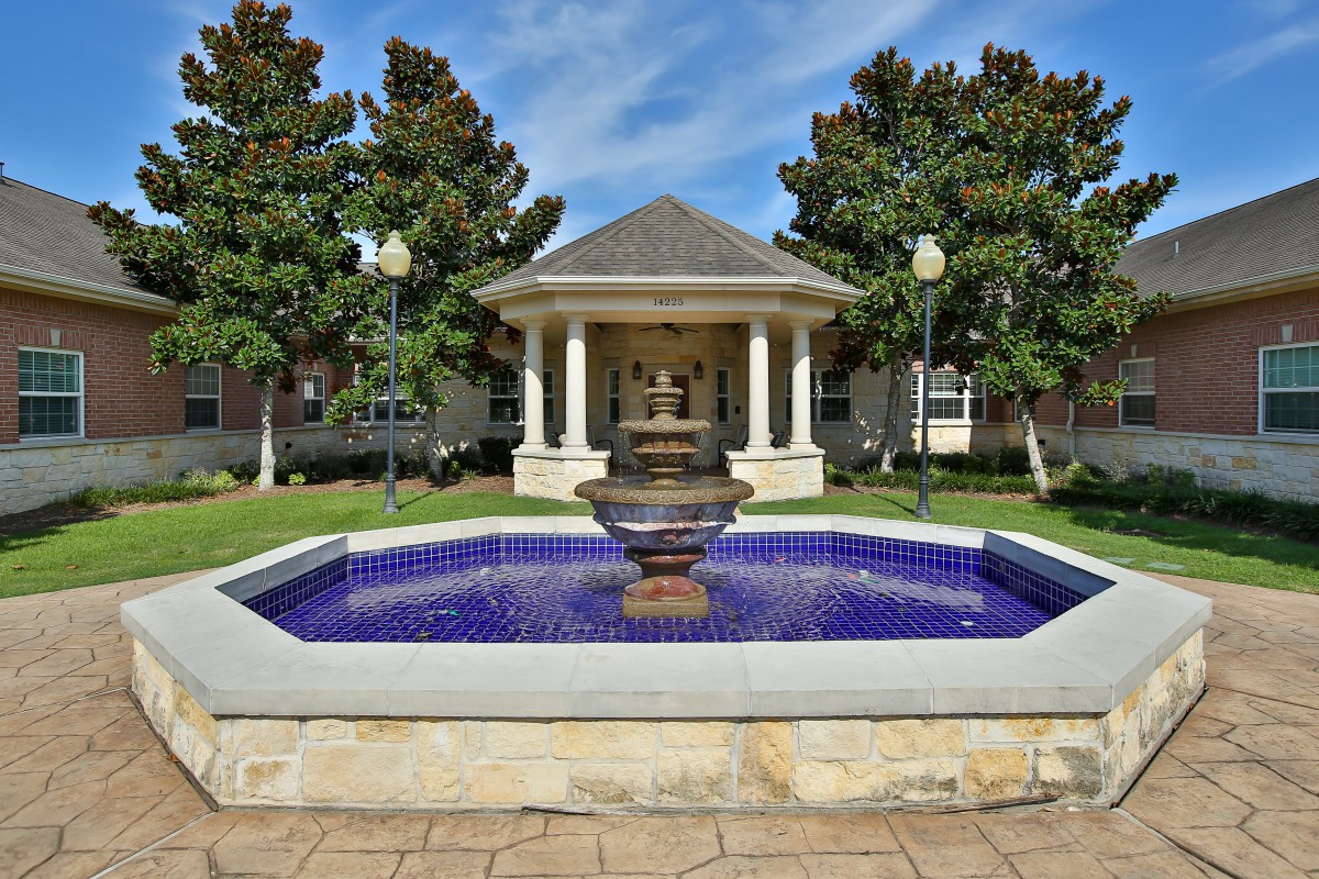 A scenic view of a fountain in front of a gazebo, surrounded by trees and greenery.