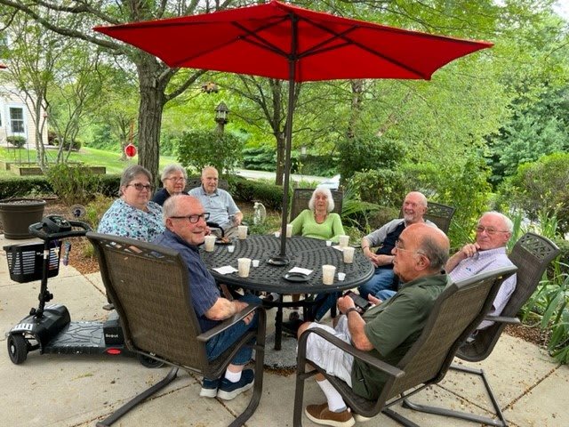A group of elderly people sitting around a patio table having a conversation under a red umbrella.