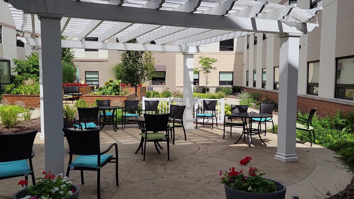 A sunny outdoor patio with tables and chairs under a pergola, surrounded by greenery.