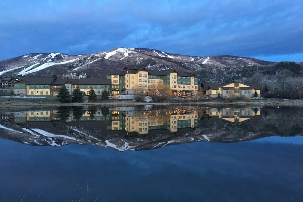 Scenic view of buildings and snowy mountains reflected in a lake at twilight.
