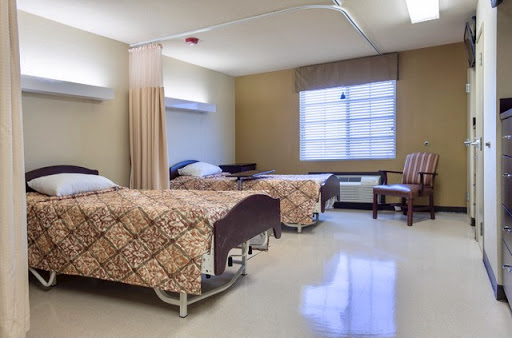 A spacious hospital patient room featuring two beds, a chair, and natural light from a window.