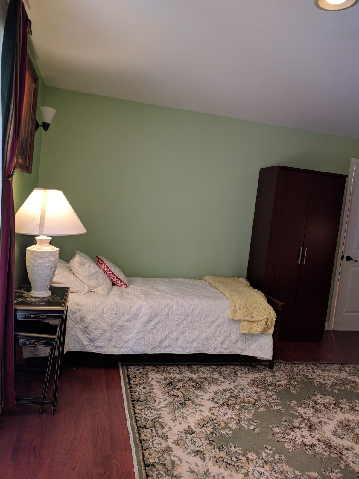 A cozy bedroom with a single bed, nightstand and lamp, against a green wall.