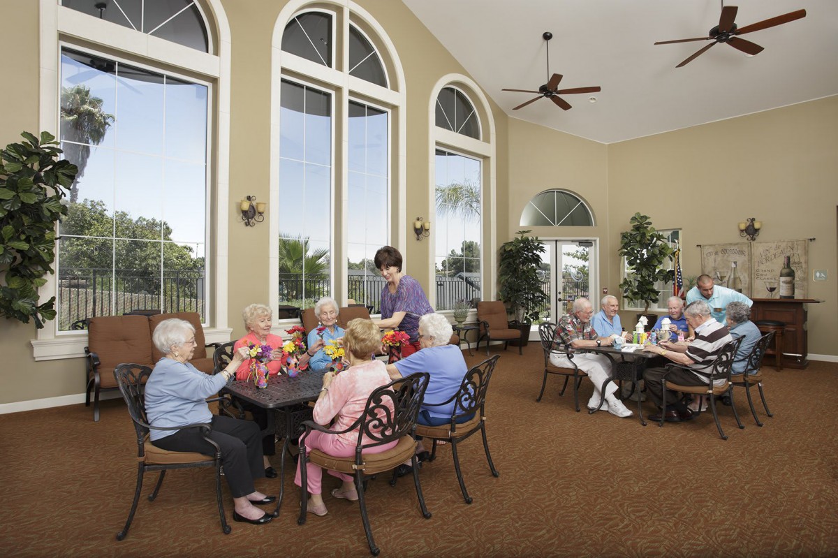 A group of seniors enjoying a social meal in a well-lit dining area with large arched windows.