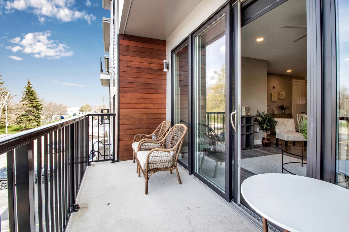 A modern balcony with outdoor seating, featuring large glass doors and a view of the sky.