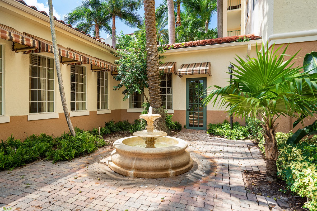 A serene courtyard featuring a central fountain surrounded by tropical plants and palm trees with architectural elements in the background.